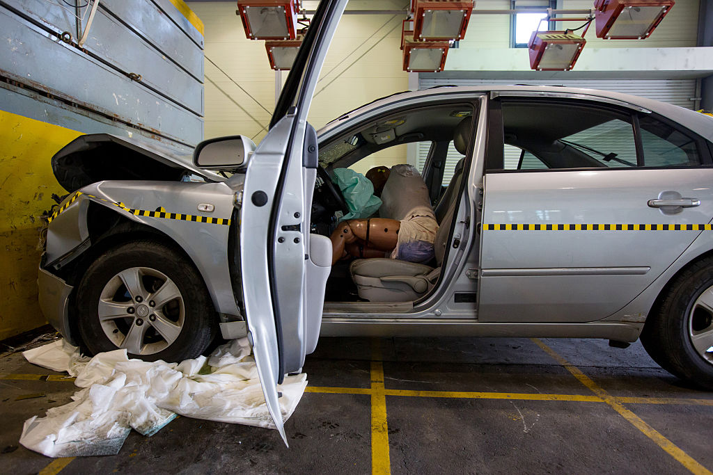 A crash test dummy sits behind a deployed airbag inside a damaged Hyundai Sonata