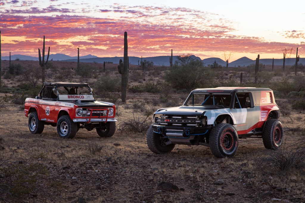The red-and-white 1969 Ford Bronco Baja racer next to the 2019 gray-and-red Ford Bronco R race prototype parked in the desert