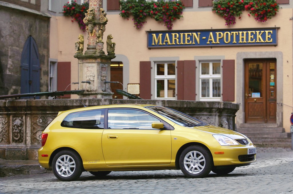 A golden yellow Honda Civic Si hatchback parked in a quaint European village.