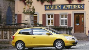 A golden yellow Honda Civic Si hatchback parked in a quaint European village.