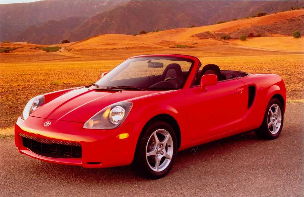 A red Toyota MR2 Spyder parked next to a picturesque countryside.
