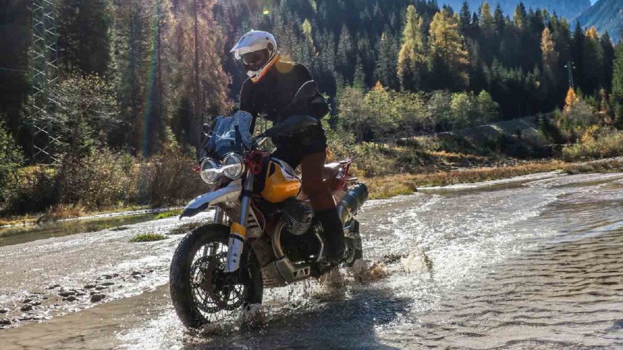 Yellow-trimmed 2020 Moto Guzzi V85TT Adventure being ridden through a mountain forest stream