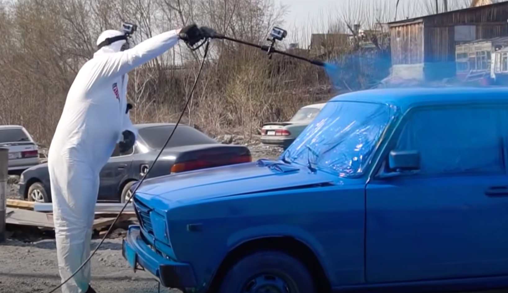 Man using pressure washer to paint Lada blue in open parking lot