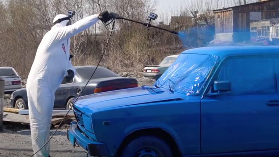 Man using pressure washer to paint Lada blue in open parking lot