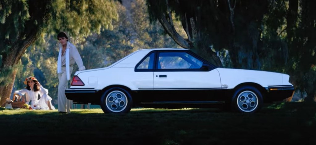 A white Ford EXP is parked in the grass as a couple have a picnic