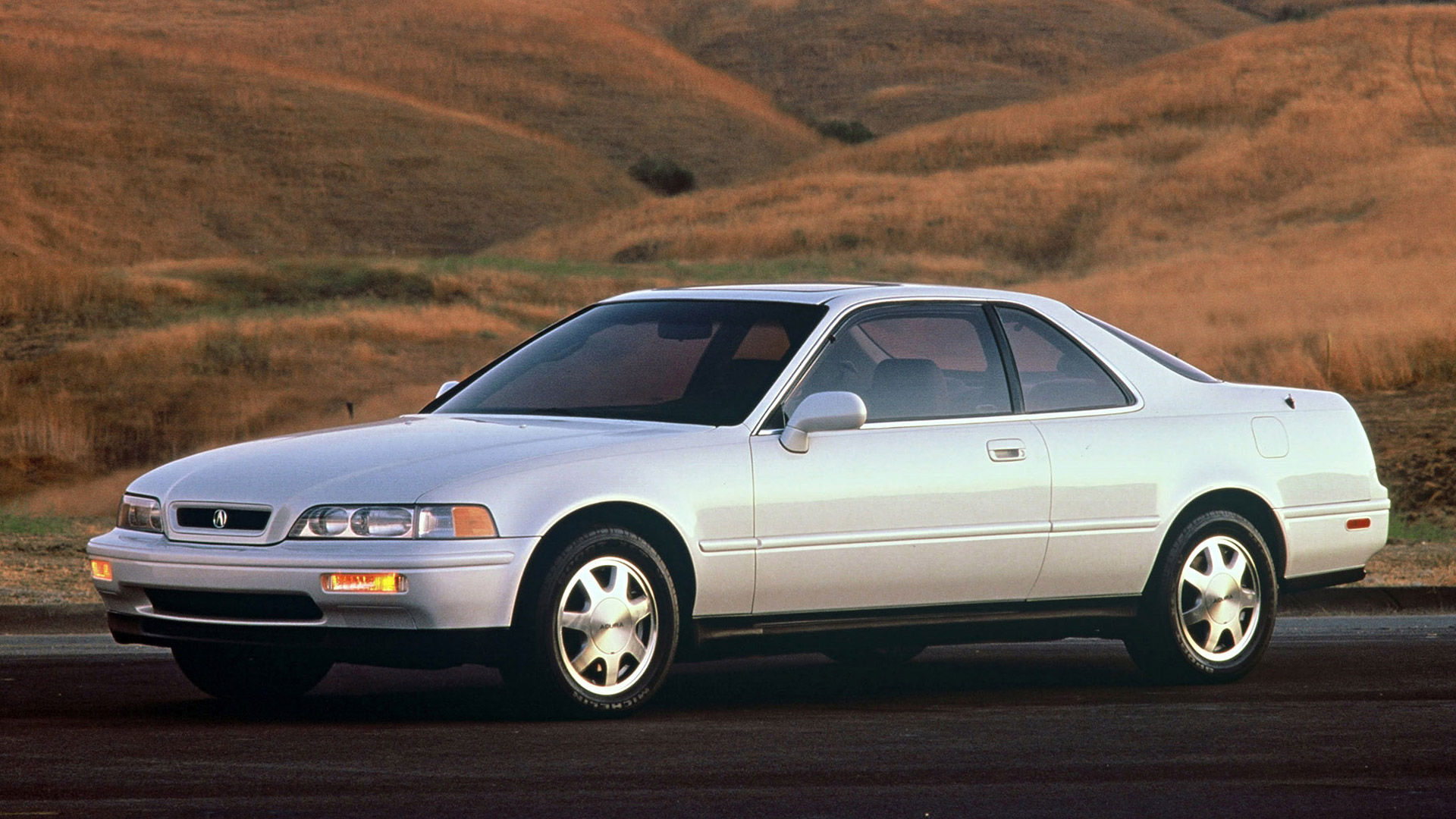 A white Acura Legend coupe sits before rolling hills.
