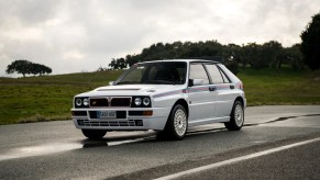 White with red-and-blue stripes 1992 Lancia Delta Integrale Evoluzione I Martini 5 Edition hot hatch on a wet road in front of a grassy hill
