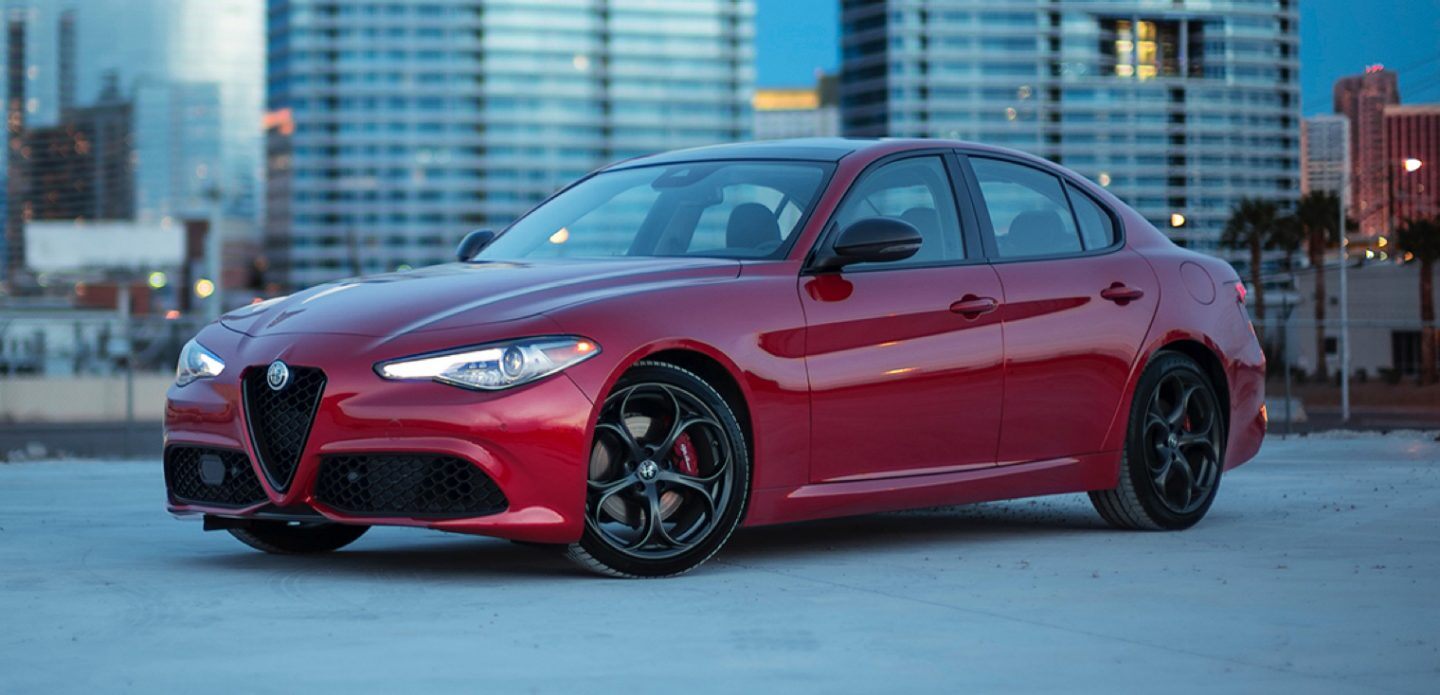 A red Alfa Romeo Giulia sedan sits in a parking lot with a city in the background.