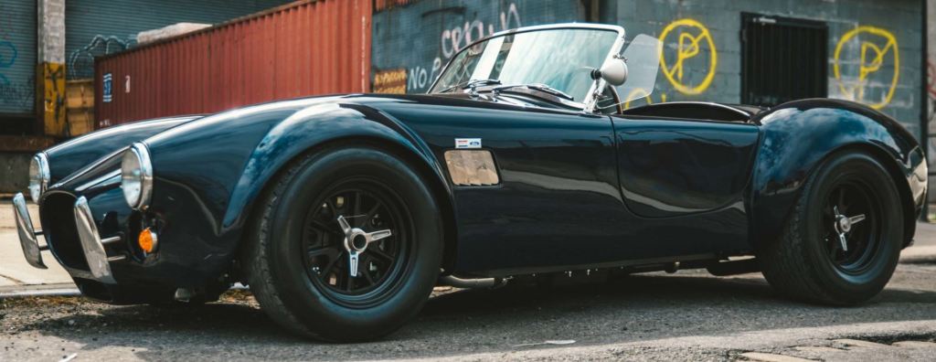 A black AC Shelby Cobra sits in an industrial area.