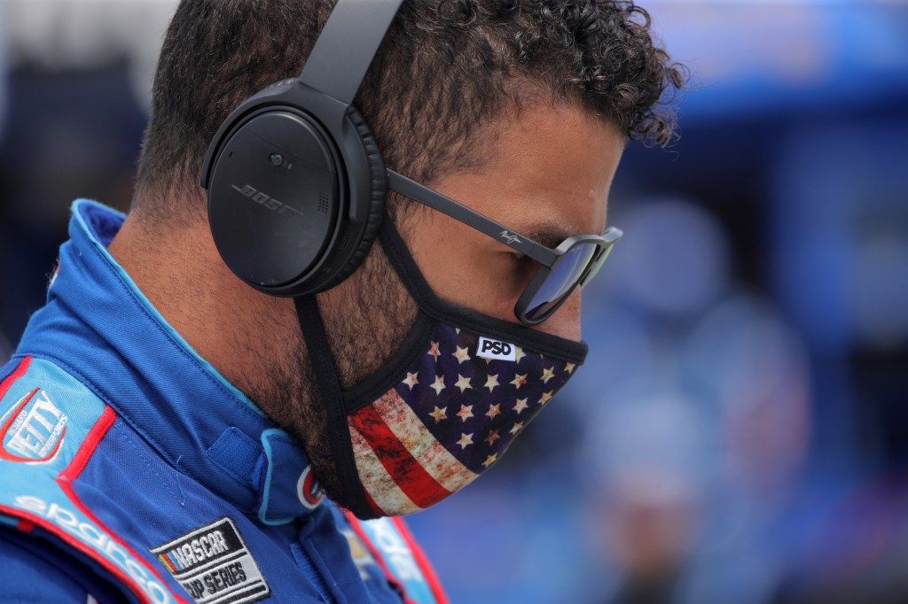 NASCAR driver Bubba Wallace stands with an American flag face mask, in his blue racing suit, with ear protection and sunglasses on