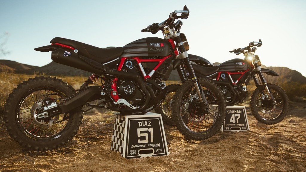 2 black-and-red Ducati Scrambler Desert Sleds modified for the Mint 400 standing on their stands in the desert
