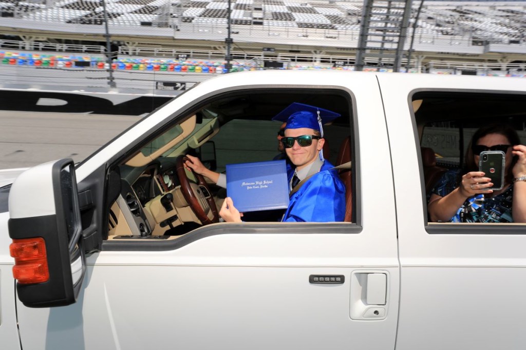 A high school senior in cap and gown shows the diploma he received at Daytona International Speedway