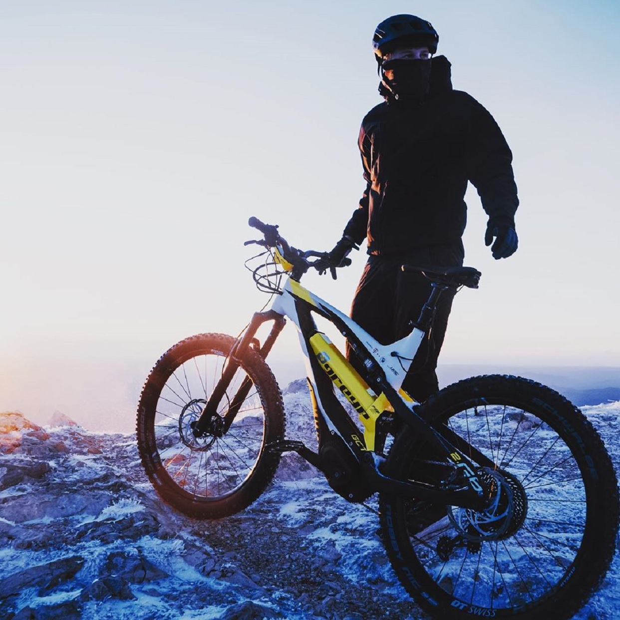 White-and-yellow Greyp G6 electric mountain bike on top of a snowy mountain facing the sunrise