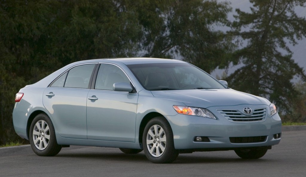 a 2007 Toyota Camry was a great car new, and its still great used. This light blue one is parked on the street in front of some gorgeous trees.