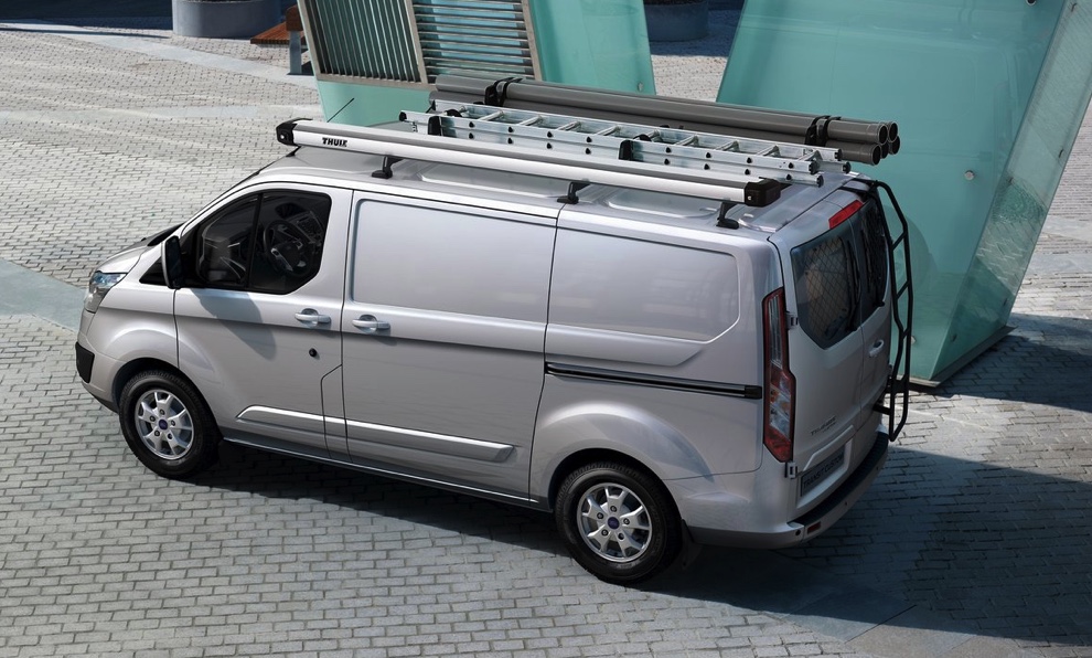 A gray Ford Transit with a custom roof mount driving through the city