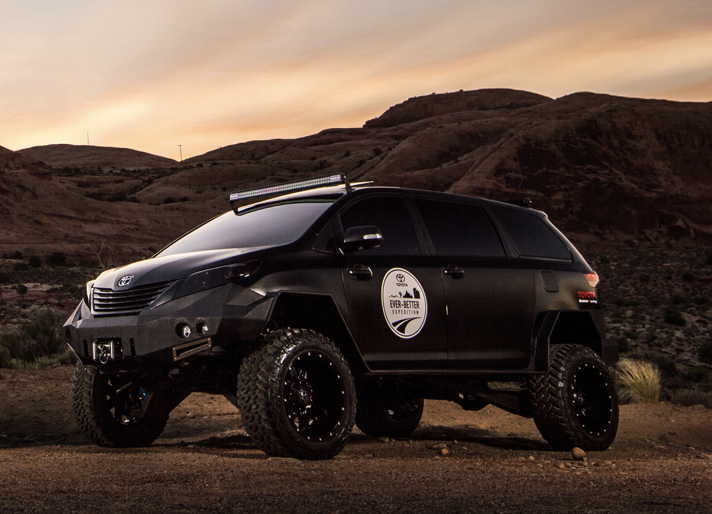 a black on black Toyota Sienna overland vehicle with massive tires in a desert scene