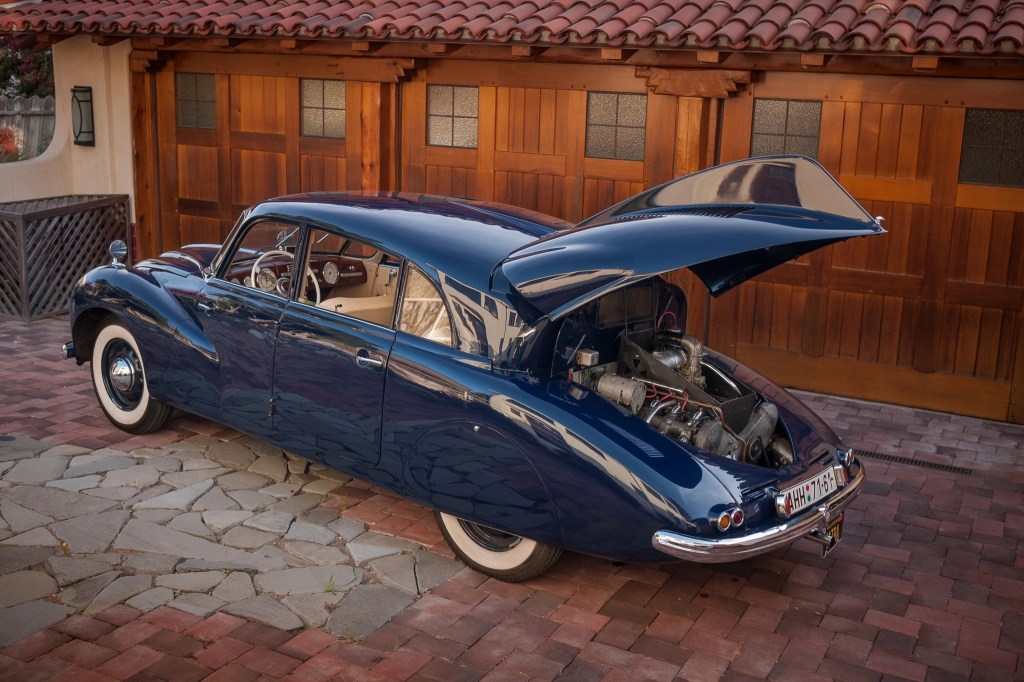 A blue Tatra T87 with its rear engine cover open in front of a wooden garage door