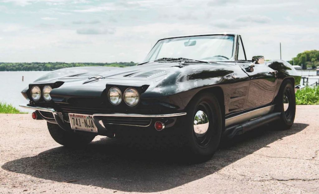 A black convertible 1963 Corvette sits in front of a river. The four flip up headlights are rotated up. 