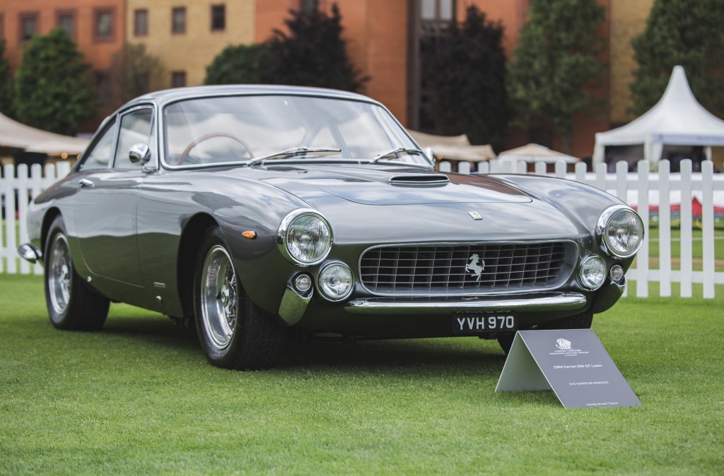 A charcoal colored 1962 Ferrari 250 GT Lusso sits on the grass on display in a car show in London.