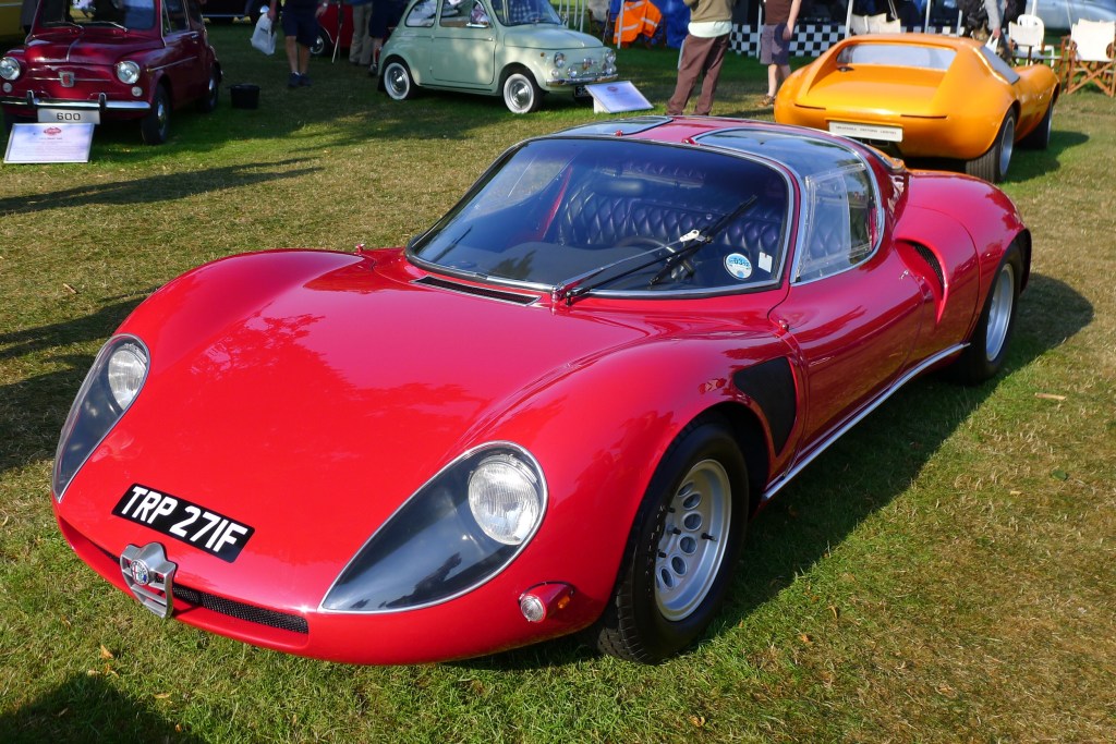 A red Alfa Romeo sports car sits on a lawn at a car show. 