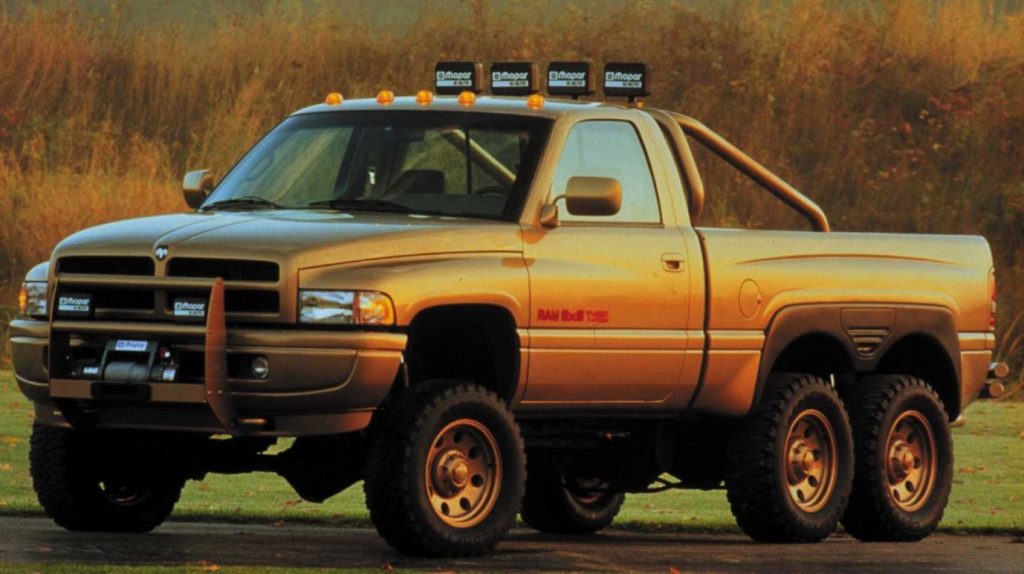 A gold colored Ram 3500 6x6 pickup sits on a road with shrubbery in the background.