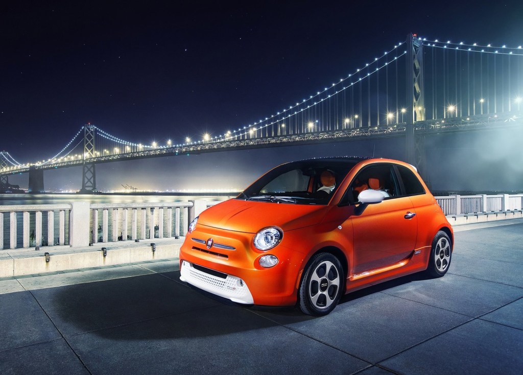 An orange 2014 Fiat 500e in front of a lit-up suspension bridge at night