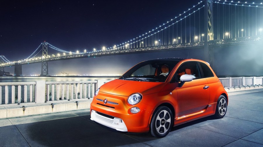 orange Fiat 500e in front of a lit suspension bridge late at night in the city.
