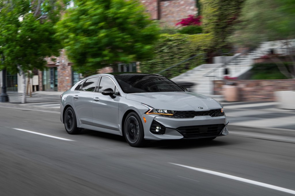A gray 2021 Kia K5 sedan passes through a downtown city street.
