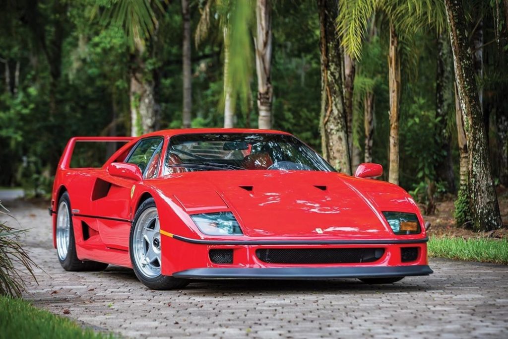 A red Ferrari F40 parked on a stone driveway in front of some palm trees