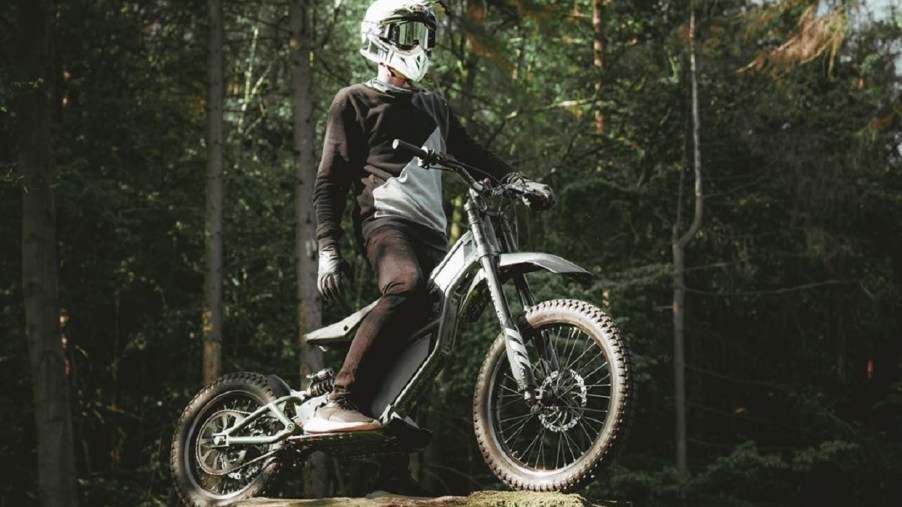 A white-helmeted rider on a green Kuberg Ranger, standing on a boulder in a forest