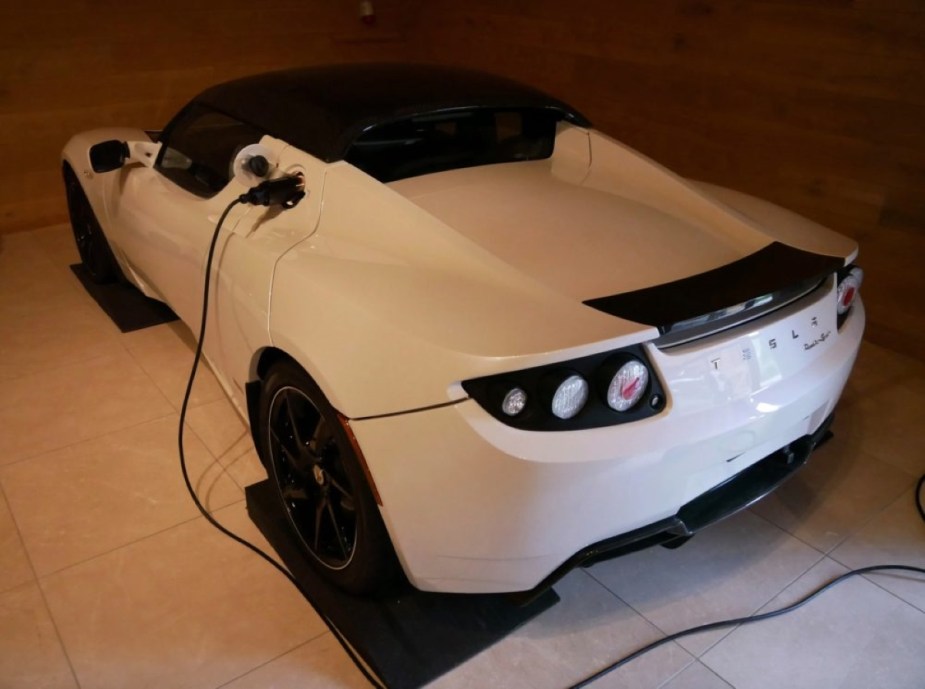 The rear of a white Tesla Roadster sits in a garage hooked up to a charging cable.