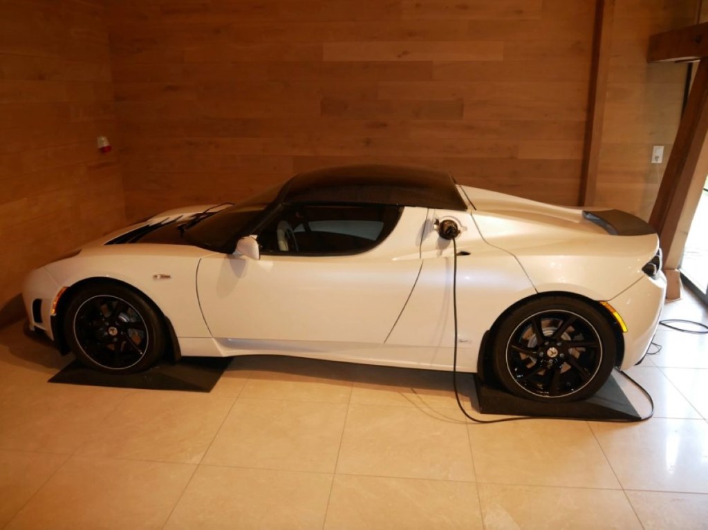 A white Tesla Roadster sits in a garage hooked up to a charging cable.