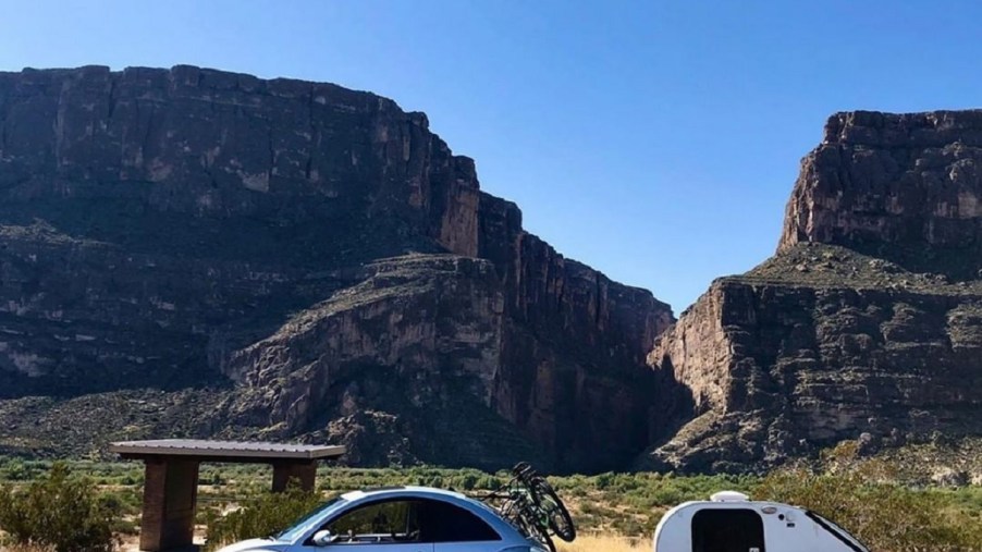A silver Volkswagen New Beetle towing a trailer, parked in front of a mountain range