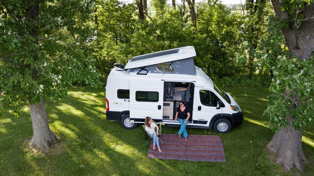 A drone takes a picture of a Winnebago Solis RV parked and set up at a campsite with two ladies sitting by it. The pop-top is extended on the roof of the RV. 