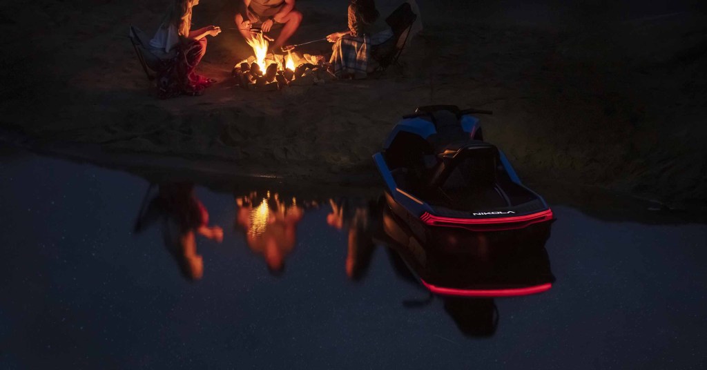 The rear lights of the Nikola Wav are on as it sits on shore by a campfire.
