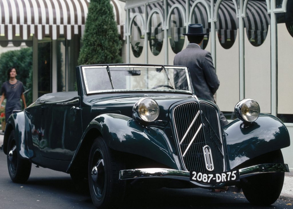 A man stands near a black 1938 Citroen Traction Avant 11B Cabriolet parked by a hotel