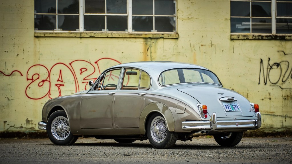 A rear-3/4 view of a two-tone gray 1964 Jaguar Mk2 in front of a yellow graffiti'd wall