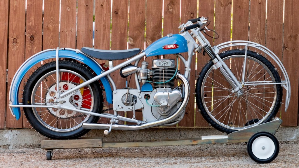 A blue 1967 Jawa/Eso factory ice racer motorcycle on a stand, with spiked ice tires in front of a wooden fence