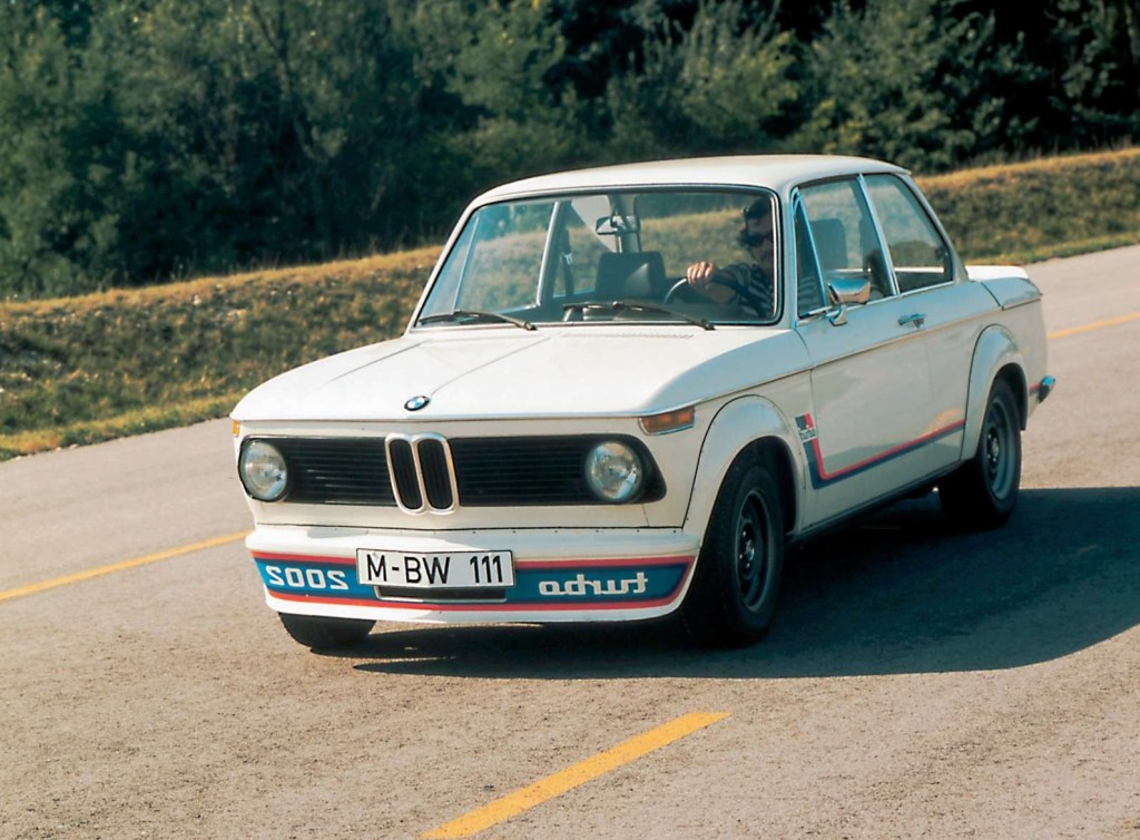 A white-with-red-and-blue-striping 1973 BMW 2002 Turbo drives down a sunny road