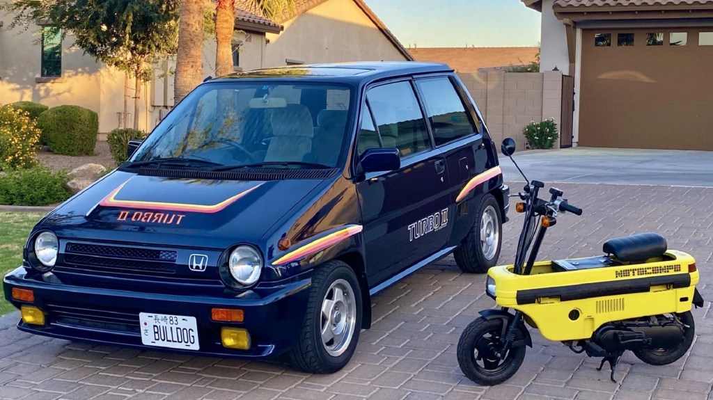 A blue 1983 Honda City Turbo II with a yellow unfolded Honda Motocompo scooter on a brick driveway