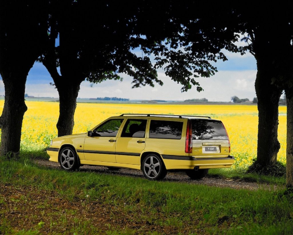 The side-rear view of a yellow 1995 Volvo 850 T-5R Wagon on a shaded dirt road by a yellow field