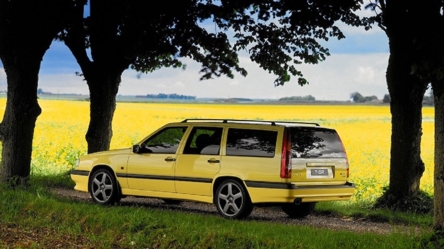 The side-rear view of a yellow 1995 Volvo 850 T-5R Wagon on a shaded dirt road by a yellow field