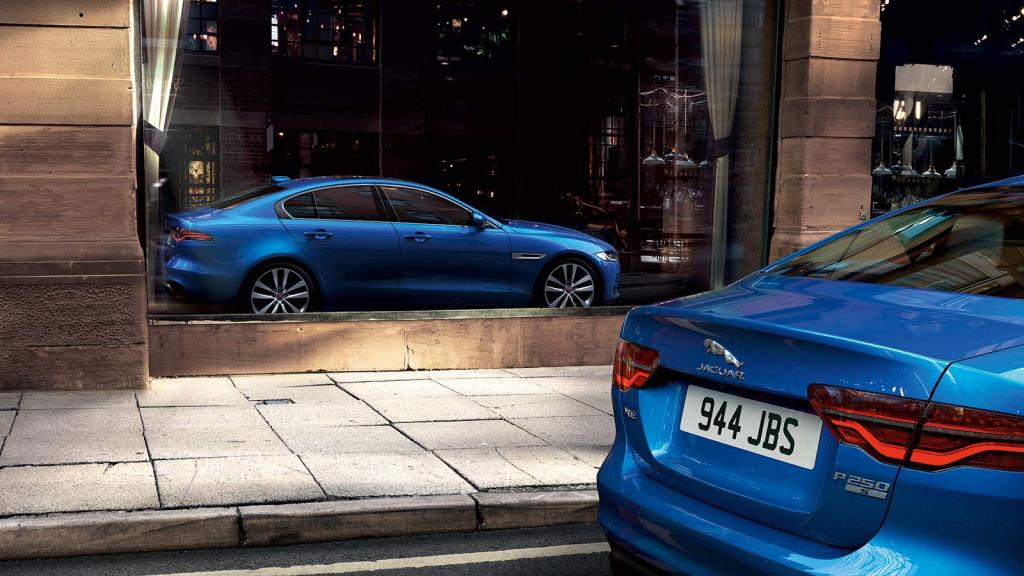 A blue 2020 Jaguar XE P 250S sedan is parked in front of a storefront. The reflection of the sedan is seen in full on the glass of the establishment.