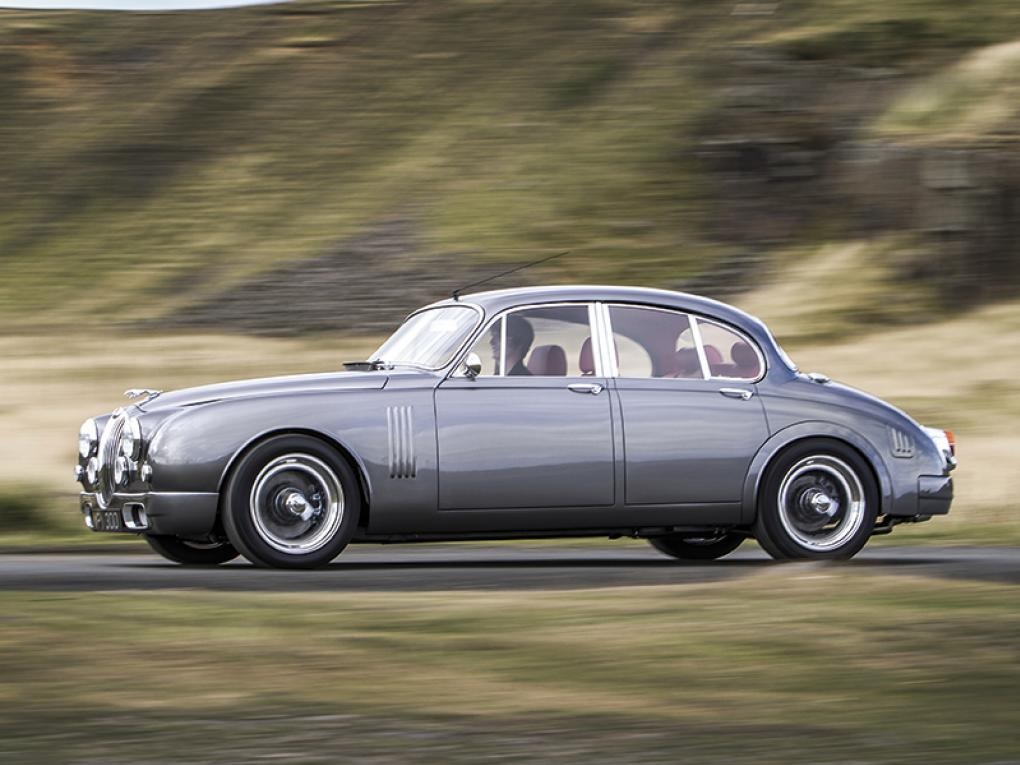A silver CMC Jaguar Mk2 by Callum drives through the British countryside
