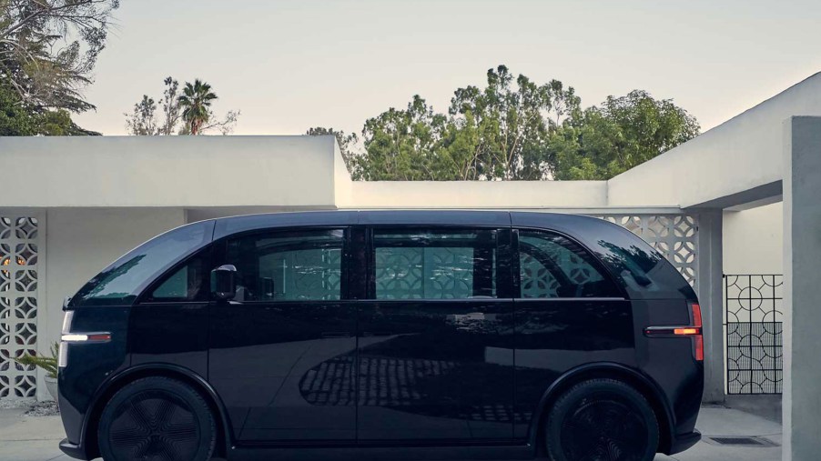 A black Canoo electric van sits in a driveway of a modern home.