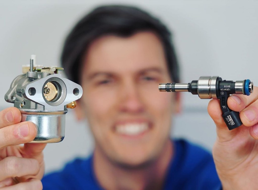 Jason Fenske of Engineering Explained holds a carburetor in his right hand and an electronic fuel injector in his left hand