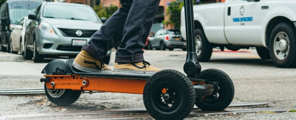 A black and orange electric scooter is shown with somebody riding it in an urban setting.