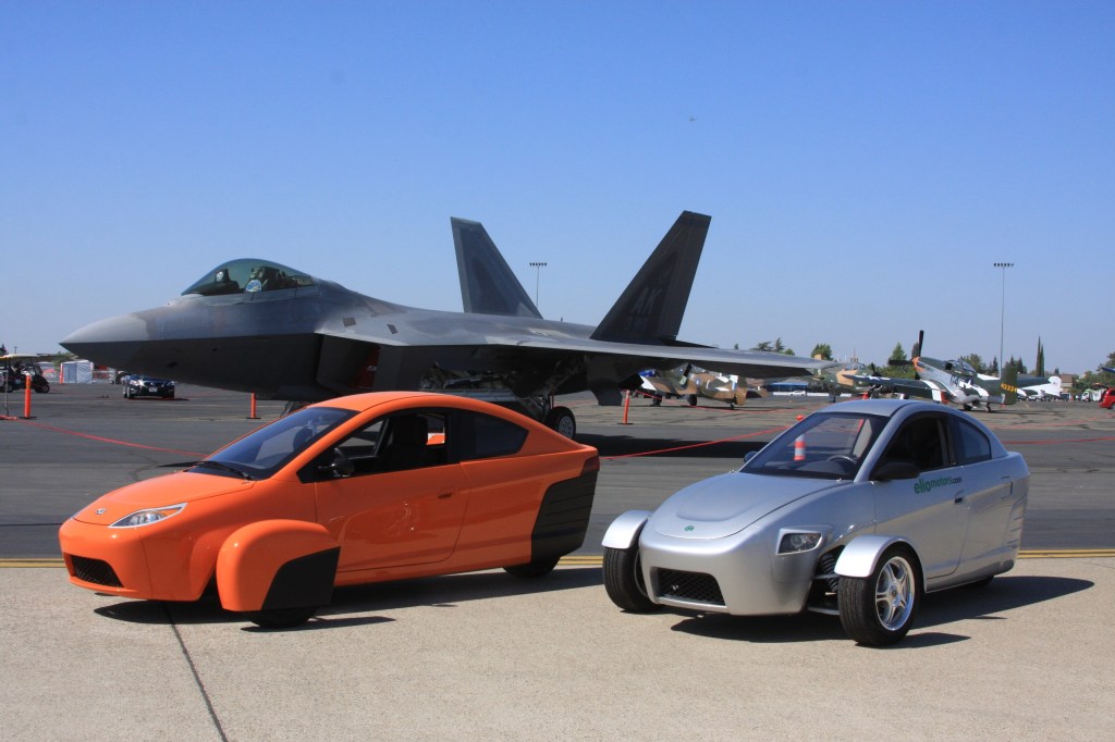 Two Elio Protypes sit on a tarmac before a parked fighter plane.