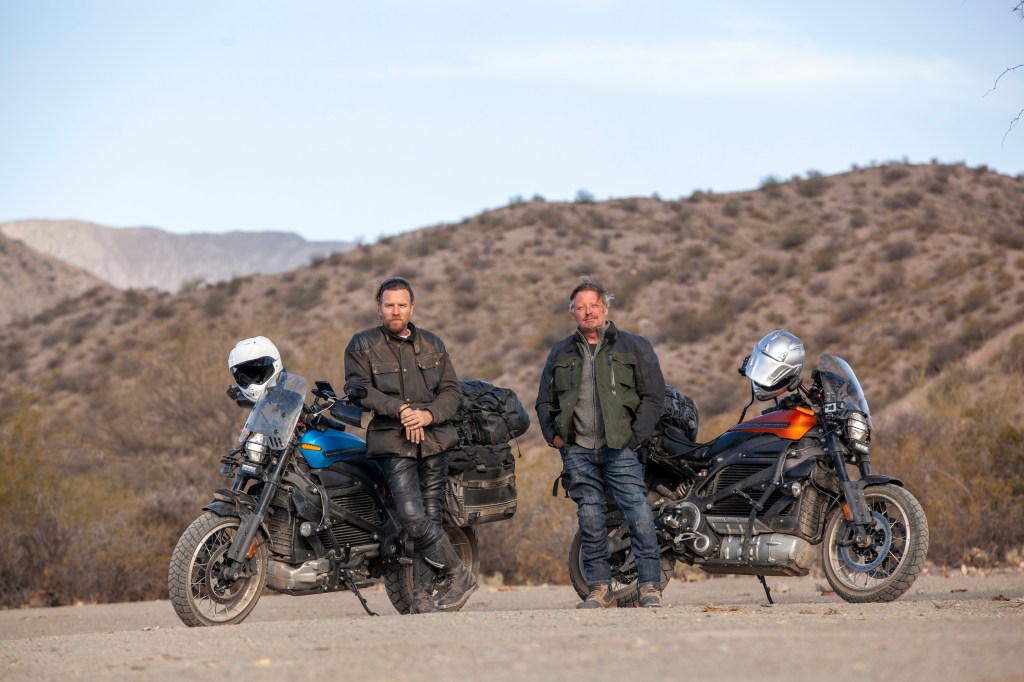 Ewan McGregor and Charley Boorman standing in front of their blue and orange Harley-Davidson LiveWires in the middle of the desert
