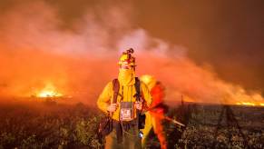 A firefighter in wildfire gear and camera in tow with a raging forest fire in the background